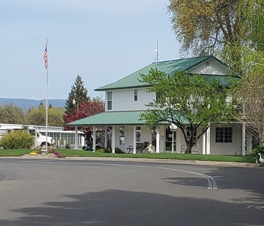 White building with a green roof, surrounded by trees and a garden. An American flag on a pole stands to the left. Overcast sky. Paved driveway in the foreground.